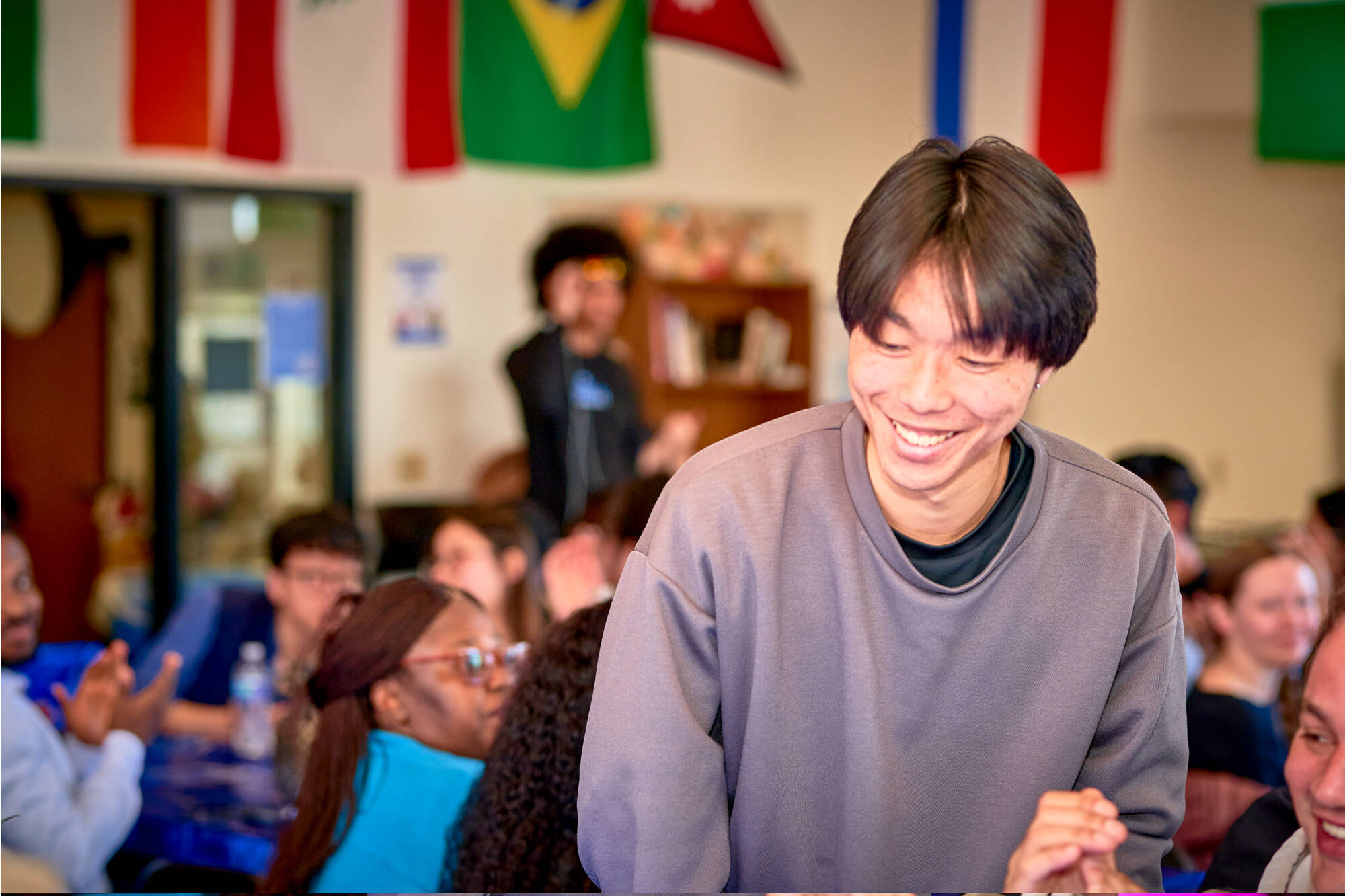student smiling in a grey sweatshirt with various country flags hanging on the wall in the back
